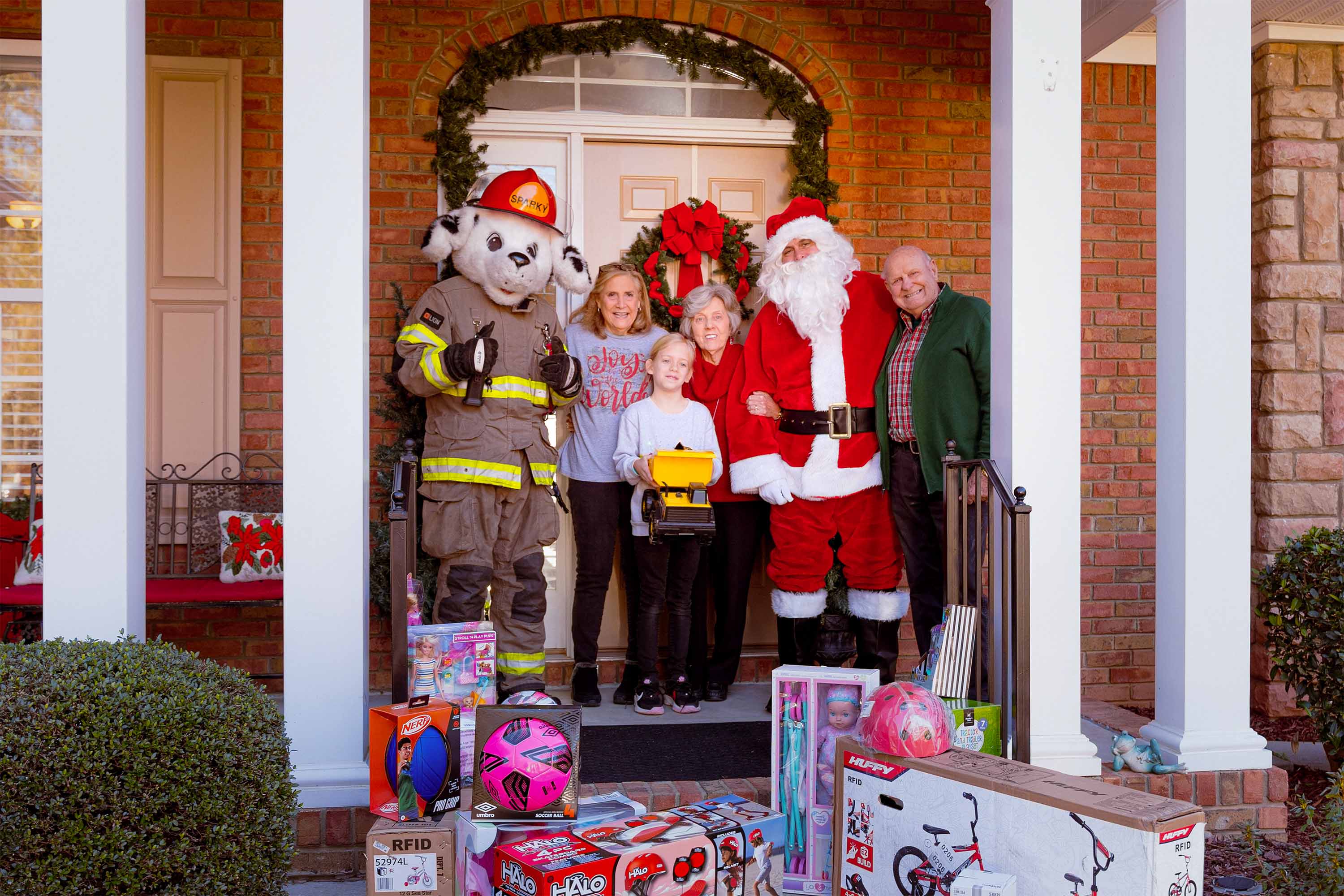 Family posing in front of their house, holding Christmas presents they are donating, with Santa and Sparky with the fire department
